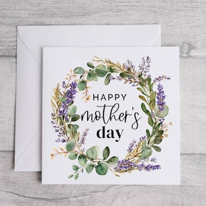 May include: A white Mother's Day card with a floral wreath design. The wreath features green eucalyptus leaves, purple lavender, and yellow accents. The words "Happy Mother's Day" are written in black script.