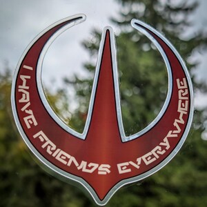 Può includere: Un adesivo rosso e argento con la scritta bianca "I HAVE FRIENDS EVERYWHERE". L'adesivo presenta un design stilizzato unico con una forma a freccia centrale ed elementi curvi. Lo sfondo è sfocato, mostrando fogliame verde.