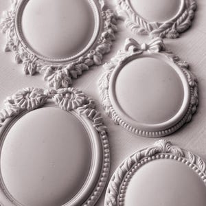 May include: Five white ornate oval picture frames with decorative edges. The frames are arranged in a cluster on a white background.