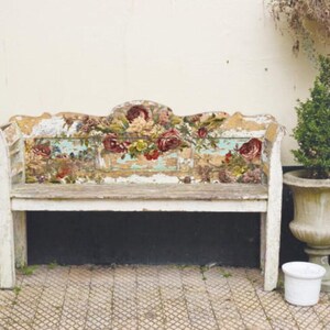 Pu&ograve; includere: Una panca di legno bianco con schienale e seduta a motivi floreali. La panca &egrave; usurata e ha un aspetto rustico. &Egrave; posizionata davanti a un muro bianco con una pianta in vaso a destra.
