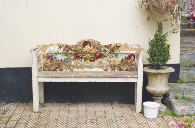 Pu&ograve; includere: Una panca di legno bianco con schienale e seduta a motivi floreali. La panca &egrave; usurata e ha un aspetto rustico. &Egrave; posizionata davanti a un muro bianco con una pianta in vaso a destra.