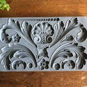 May include: Gray rectangular mold with an ornate floral and leaf design. The mold features detailed carvings of stylized flowers, leaves, and scrollwork, perfect for creating decorative elements. The mold is made of a flexible material.