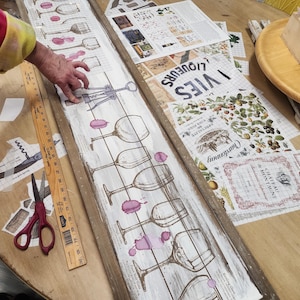 Pu&ograve; includere: Un cartello di legno bianco con una fila di bicchieri da vino e un cavatappi. Il cartello &egrave; decorato con una macchia marrone chiaro e ha il testo "VIES" e "LIQUEURS" stampato su di esso.