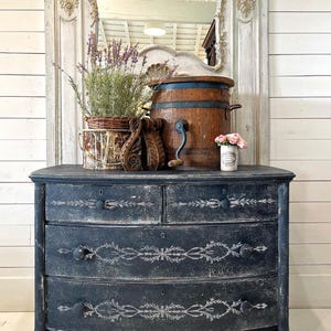 Pu&ograve; includere: Un antico com&ograve; dipinto di nero con tre cassetti e un fronte curvo. I cassetti presentano un motivo a stencil decorativo in vernice bianca. Il com&ograve; &egrave; appoggiato su un pavimento di legno.