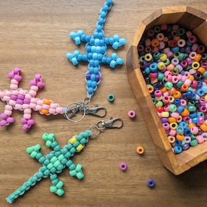 May include: Three beaded lizard keychains in pink, blue, and green, with silver clasps, are displayed on a wooden surface. A heart-shaped wooden bowl filled with colourful beads is also present. The keychains are made of small, colourful beads.