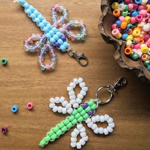 Op de afbeelding: Twee libel-vormige sleutelhangers gemaakt van kleurrijke plastic kralen. De ene heeft lichtblauwe en iriserende kralen met een metalen karabijnhaak, de andere heeft limoengroene, paarse en witte kralen met een metalen ring en sluiting.