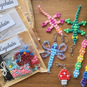 Könnte beinhalten: Eine Sammlung farbenfroher Schlüsselanhänger aus Perlen in verschiedenen Formen, darunter Eidechsen, ein Schmetterling und ein Pilz. DIY-Schlüsselanhänger-Kits sind sichtbar, mit dem Text "DIY Keychain" auf der Verpackung. Die Schlüsselanhänger sind an silbernen Verschlüssen befestigt.