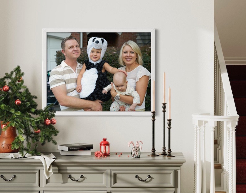 May include: A rectangular white picture frame containing a photograph of a family of four. The photo shows a man, a woman, a toddler wearing a black and white panda costume, and an infant. The frame is displayed on a gray wooden console table.