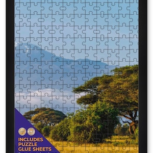 May include: A rectangular jigsaw puzzle featuring a landscape of African acacia trees and a distant mountain under a clear blue sky. The puzzle is enclosed in a black frame and includes a purple corner label stating INCLUDES PUZZLE GLUE SHEETS.