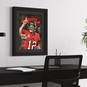 May include: Framed Sports Illustrated magazine cover featuring Tom Brady in a red Tampa Bay Buccaneers jersey. The cover reads "THE GOAT Says Goodbye" and is displayed on a white wall above a black desk.