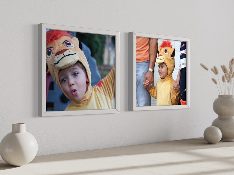 May include: Two square white frames mounted on a wall, each displaying a photograph of a child wearing a yellow lion costume with a bright red mane. The frames are positioned above a surface featuring a round white vase and a stacked ceramic vase.