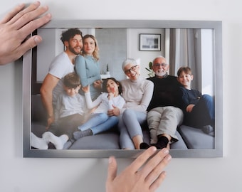 May include: A rectangular silver-toned metal picture frame containing a color photograph of a multi-generational family sitting together on a gray sofa. The frame has a clean, flat profile and is being held against a plain white wall.