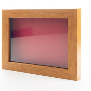 May include: A wooden picture frame with a red mat and a clear glass front. The frame is a light brown color and has a simple design.