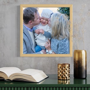May include: A framed photograph of a family with a gold frame. The photo shows a man and woman kissing a baby on the cheek. The baby is wearing a white knitted hat.