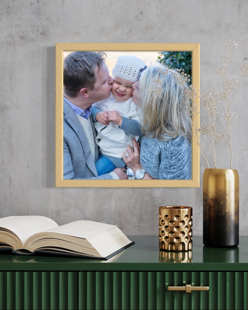 May include: A framed photograph of a family with a gold frame. The photo shows a man and woman kissing a baby on the cheek. The baby is wearing a white knitted hat.