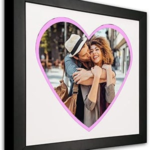 May include: A black picture frame with a white mat and a pink heart-shaped cutout. The cutout features a couple embracing, with the man kissing the woman on the cheek.