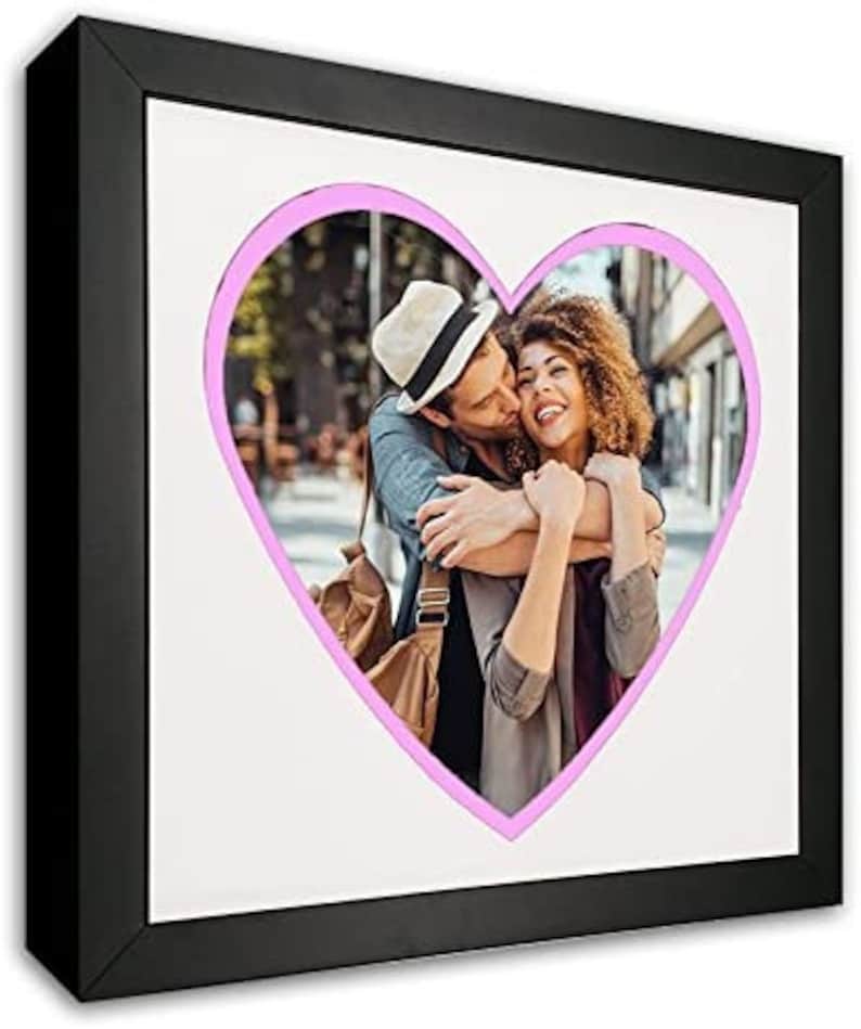 May include: A black picture frame with a white mat and a pink heart-shaped cutout. The cutout features a couple embracing, with the man kissing the woman on the cheek.