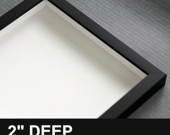 May include: A square black shadow box frame with a white interior backing. The frame features a 5.08 cm deep profile. The text 2" DEEP is printed at the bottom of the image, indicating the depth of the frame construction.