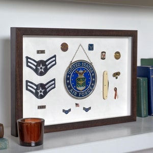 May include: A rectangular dark wood shadow box frame containing a collection of United States Air Force memorabilia. The display features two blue and white chevron rank patches, a circular United States Air Force emblem, pins, and a small pocket knife.
