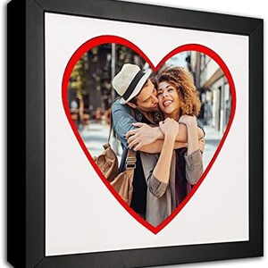 May include: A black picture frame with a red heart-shaped cutout. The cutout features a couple embracing in a city street.