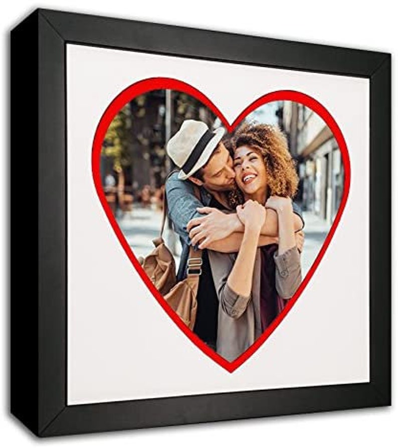 May include: A black picture frame with a red heart-shaped cutout. The cutout features a couple embracing in a city street.