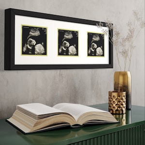 May include: A black picture frame with three white mats and gold trim holds three ultrasound images of a baby. The frame is hanging on a gray wall above a green table with a book and gold decorative items.