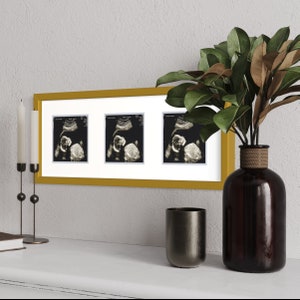 May include: A gold-colored picture frame with three black and white ultrasound images of a baby. The frame is on a white surface with a brown vase and a silver cup.
