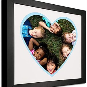 May include: A black picture frame with a white mat and a blue heart-shaped cutout. The cutout shows five children lying on their backs in a grassy field, smiling and looking at the camera.