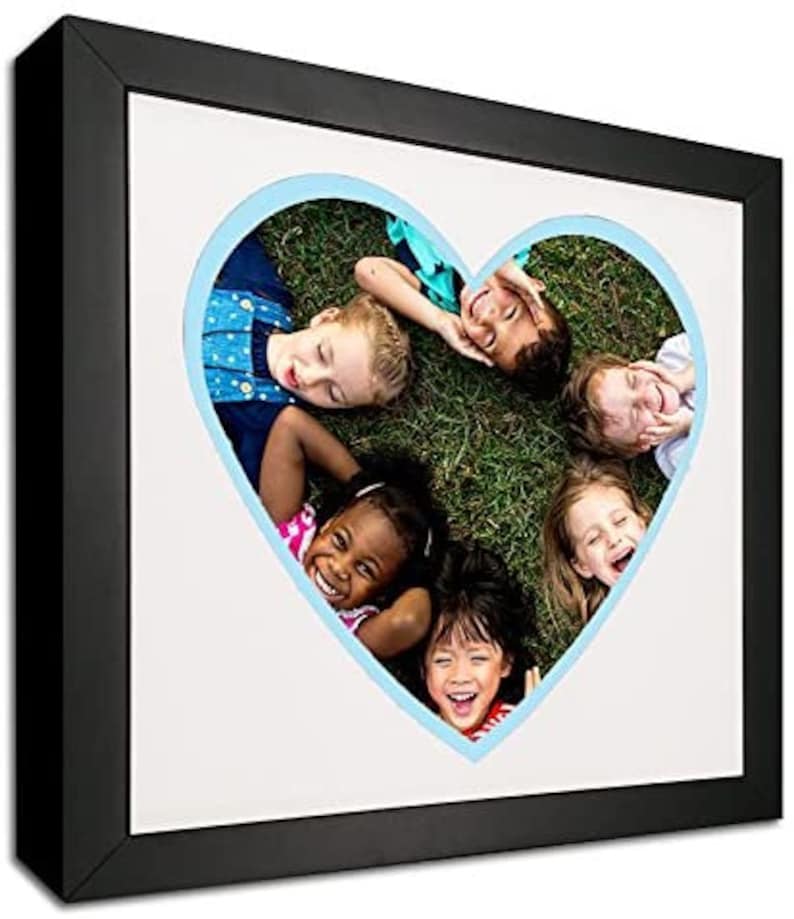 May include: A black picture frame with a white mat and a blue heart-shaped cutout. The cutout shows five children lying on their backs in a grassy field, smiling and looking at the camera.