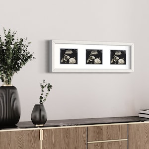 May include: A white framed print featuring three ultrasound images of a fetus. The print is displayed above a wooden cabinet with black vases and greenery. The cabinet has gold accents and a marble top.