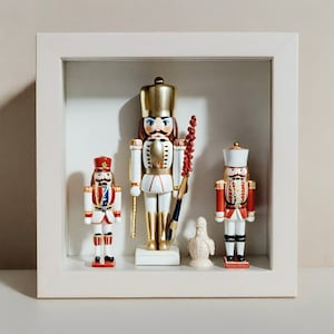 May include: A white square shadow box frame containing three nutcracker figurines and one small white bird figurine. The central nutcracker is tall with gold accents, while the two flanking nutcrackers feature red, white, and blue details.