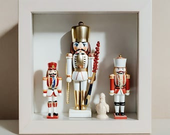 May include: A white square shadow box frame containing three nutcracker figurines and one small white bird figurine. The central nutcracker is tall with gold accents, while the two flanking nutcrackers feature red, white, and blue details.
