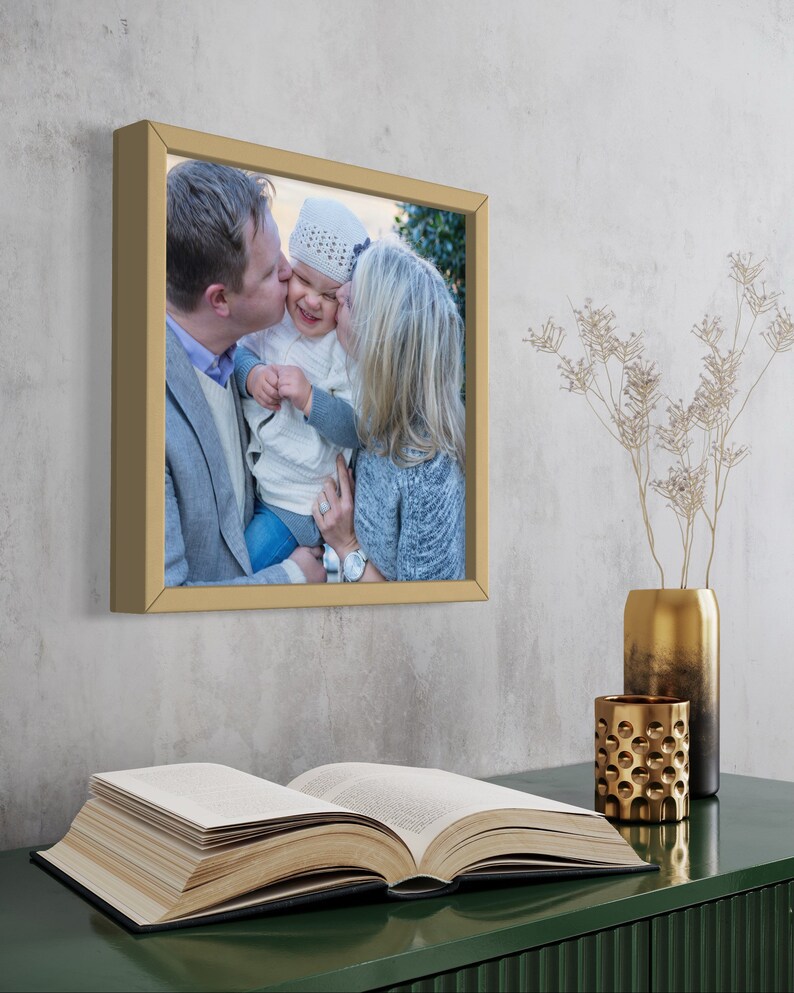 May include: A framed photograph of a family with a gold frame. The photo shows a man kissing a baby on the cheek while a woman holds the baby. The photo is on a green surface with a book and gold decorative items.