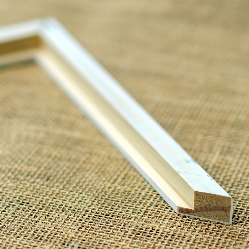 May include: An L-shaped corner section of a picture frame featuring a smooth white finish on the outer edges and a visible light brown wood grain texture on the inner side where the frame has been cut to reveal the raw material composition.