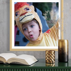 May include: A child wearing a lion costume with a surprised expression. The costume is yellow and brown with a lion's face on the head. The child is looking at the camera. The photo is framed in a light wood frame.