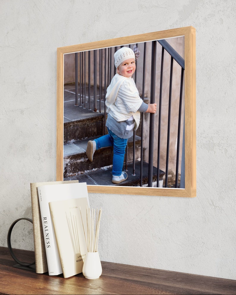 May include: A framed photograph of a young child wearing a white knitted hat and a white sweater, standing on a set of stairs and holding onto a metal railing. The child is looking at the camera and smiling.