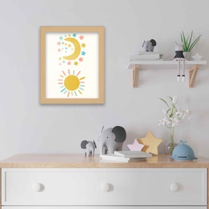 May include: A framed print with a yellow sun and a crescent moon with stars in a light blue, pink, and yellow color scheme. The print is hanging on a white wall above a wooden dresser with a drawer.