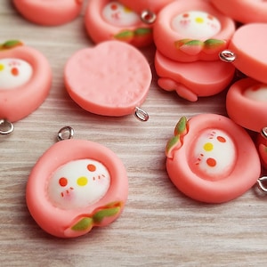 Könnte beinhalten: Eine Sammlung von pfirsichförmigen Anhängern mit einem niedlichen Comic-Gesicht und einer silbernen Schlaufe zum Aufhängen. Die Anhänger sind aus rosa Polymer Clay gefertigt und haben eine glänzende Oberfläche.
