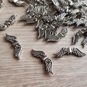 Könnte beinhalten: Ein Haufen silberner Engelsflügel-Anhänger aus Metall. Die Anhänger sind etwa 2,5 cm lang und haben ein detailliertes Design.