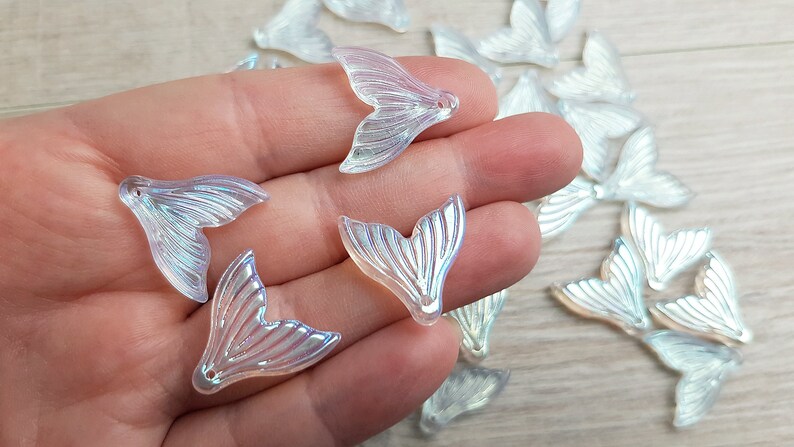 以下が含まれることがあります： 虹色のマーメイドテールのチャームのセット。透明なプラスチック製で、真珠のような光沢があります。チャームは魚の尾の形をしていて、上部に穴が開いており、ジュエリーやその他のクラフトに取り付けることができます。