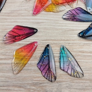 Könnte beinhalten: Sortiment aus bunten Flügeln aus Harz mit goldenen Glitzerakzenten. Die Flügel sind durchscheinend und haben ein zartes, filigranes Design.