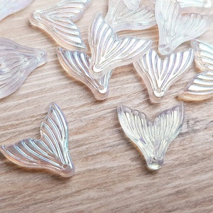 Könnte beinhalten: Eine Sammlung von regenbogenfarbenen weißen Meerjungfrauen-Schwanz-Anhängern. Die Anhänger bestehen aus klarem Kunststoff mit Perlmutt-Finish. Sie sind in Form eines Meerjungfrauen-Schwanzes mit einem gewellten Rand geformt.