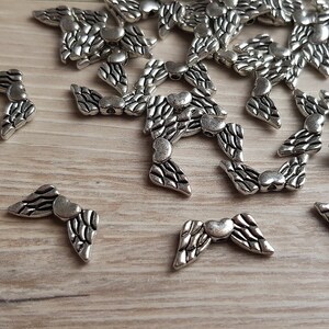 Könnte beinhalten: Ein Haufen silberner Engelsflügel aus Metall mit einem Herzen in der Mitte jedes Flügels. Die Flügel sind auf einer hellbraunen Holzoberfläche verstreut.