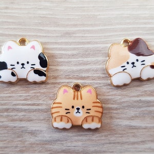 Op de afbeelding: Drie gouden metalen bedels in de vorm van katten. De eerste bedel is wit met zwarte vlekken en roze oren. De tweede bedel is wit met bruine en oranje vlekken en roze oren. De derde bedel is oranje met zwarte strepen en roze oren.