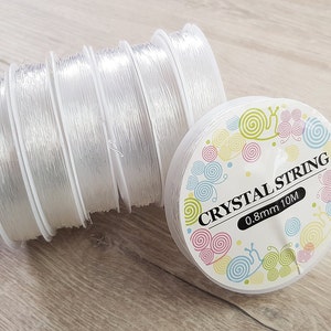 Könnte beinhalten: Eine Rolle durchsichtiges elastisches Garn zum Schmuck basteln. Das Etikett lautet "Crystal String 0.8mm 10M".
