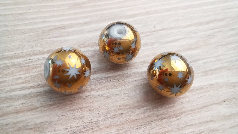Puede incluir: Tres cuentas de vidrio de color dorado con dise&ntilde;os de estrellas blancas. Las cuentas son redondas y tienen un acabado brillante.