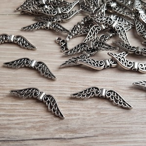 Könnte beinhalten: Eine große Menge an silbernen Engelsflügel-Anhängern aus Metall. Die Flügel sind mit filigranen Designs verziert und haben oben eine Öse zum Anbringen an Schmuck oder andere Bastelarbeiten.