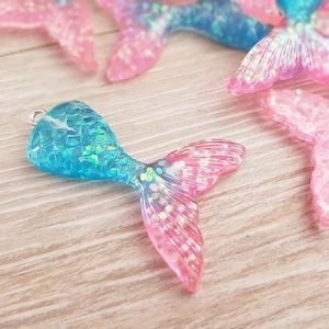 Op de afbeelding: Een blauwe en roze zeemeerminstaartbedel met glitter. De staart is gemaakt van plastic en heeft een zilveren lus om aan een ketting of armband te bevestigen.