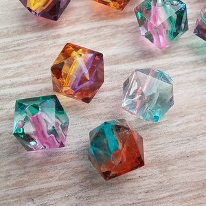 Op de afbeelding: Assortiment van kleurrijke, gefacetteerde glazen kubus kralen. De kralen zijn doorschijnend en hebben kleurwervelingen erin. De kralen liggen op een licht houten oppervlak.