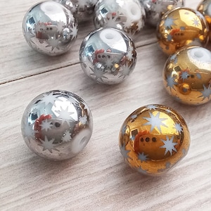 Puede incluir: Una colecci&oacute;n de adornos navide&ntilde;os plateados y dorados con un dise&ntilde;o de copo de nieve blanco. Los adornos son redondos y tienen un acabado brillante.